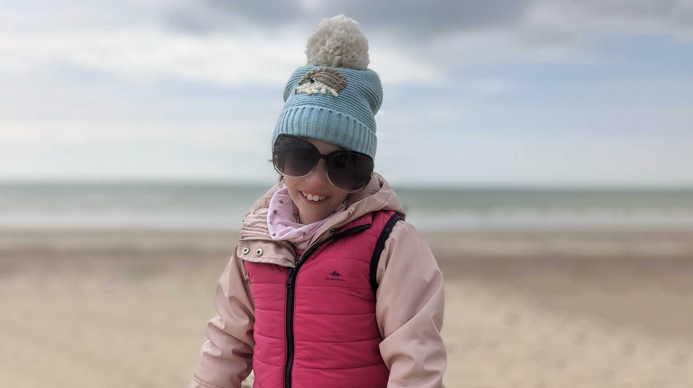 Ella wearing sunglasses and a bobble hat on the beach during her wish to have a UK coastal holiday.