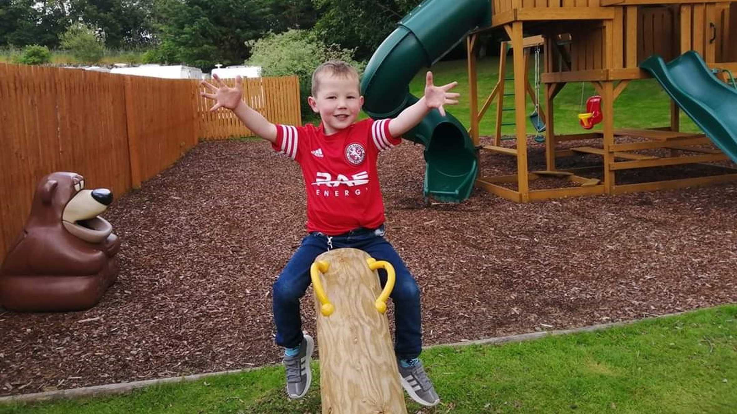 Scott, wearing a red football shirt and jeans, waving and smiling while sitting on a see-saw in a playground.